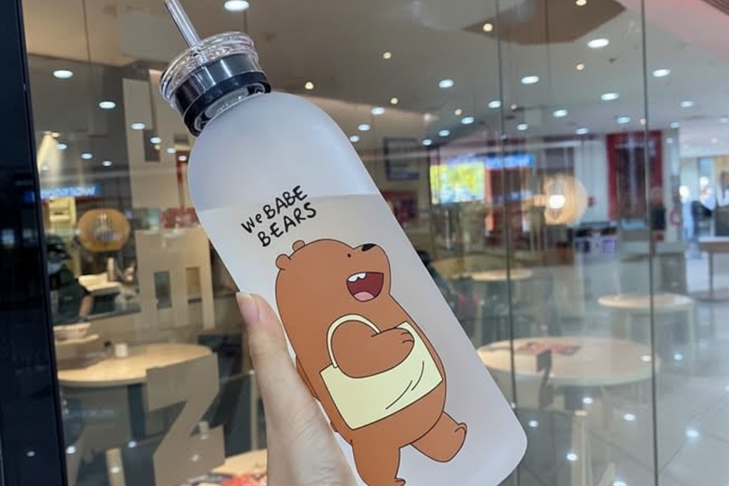 Clear water bottle with cartoon bear design held in front of a glass window.